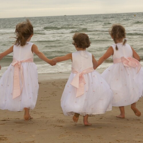 Three barefoot flower girls in white dresses with pink sashes holding hands as they run toward waves on a beachfront.