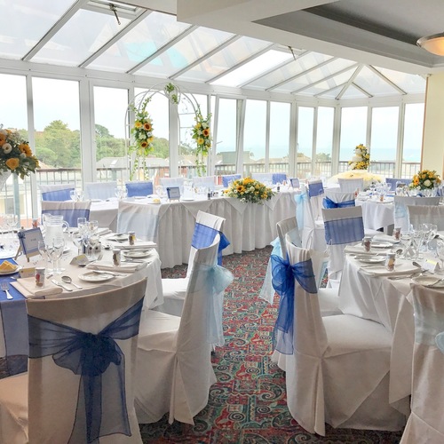 Sunlit wedding reception in a glass conservatory with linen tables, blue chair sashes, sunflower centerpieces and seaside view.