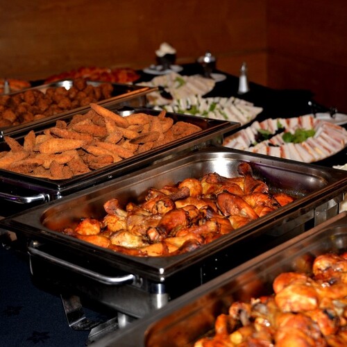 Hot buffet spread with roasted chicken drumsticks, breaded finger foods and assorted tea sandwiches in stainless chafing dishes.