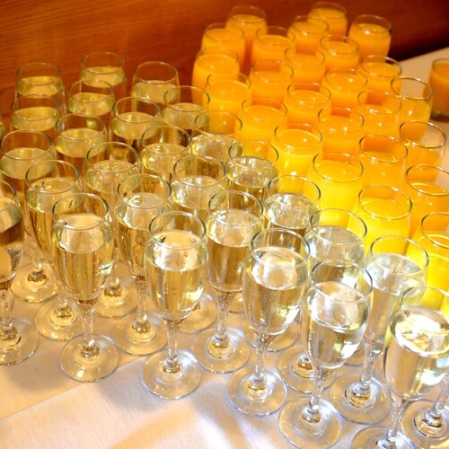 Rows of champagne flutes of sparkling wine and glasses of orange juice on a white-clothed buffet, ready for brunch