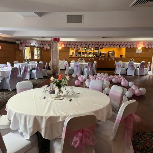 Decorated banquet room with round tables in white linen, pink chair sashes, floral centerpieces, balloon garlands and bar