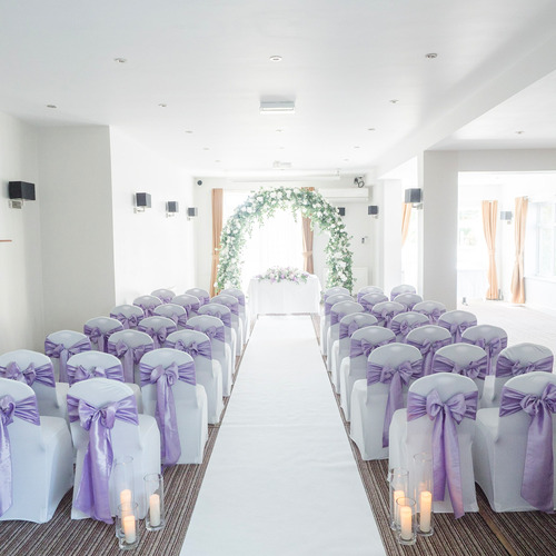 Bright ceremony room with white aisle, rows of chairs tied with lavender bows, candles and a lush floral arch.