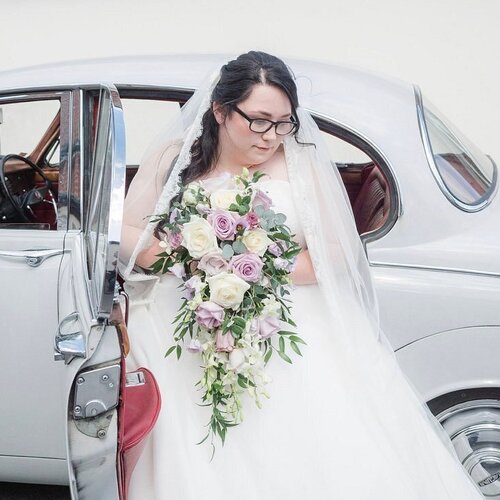 Bridal portrait stepping from silver vintage car, white gown and veil, holding cascading bouquet of white and lavender roses.