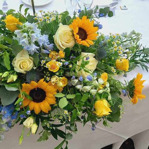 Bright sunflower and yellow-rose centerpiece on white-clothed dining table with greenery, blue accents, plates and glassware.