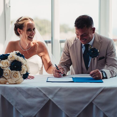 Bride in white gown and groom in beige suit sign wedding register at a linen-draped hotel table; cream and navy rose bouquet, sunlit balcony view.