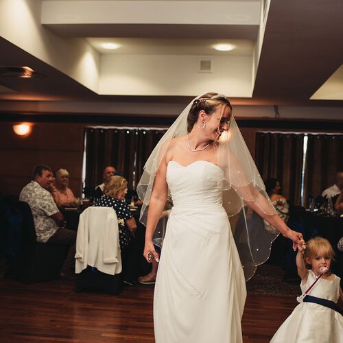 Bride in strapless gown and veil dancing with young flower girl at warm-lit hotel wedding reception with seated guests.