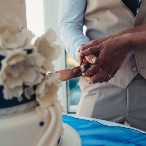 Newlyweds cutting a tiered wedding cake together, hands with wedding rings, groom in beige vest, white sugar flowers.