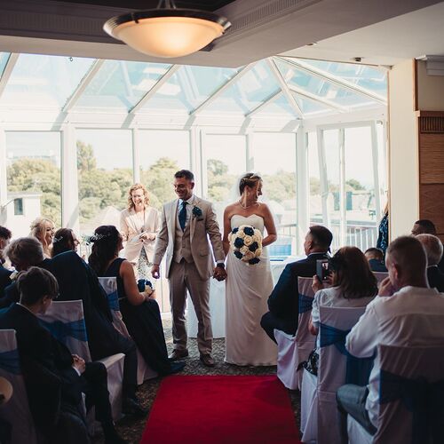 Bride and groom smile at the altar in a hotel glass-roofed function room, guests on white chairs with navy sashes.
