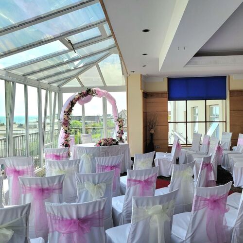 Sunlit glass conservatory set for a wedding: white chairs with pink sashes, floral arch and ocean view.