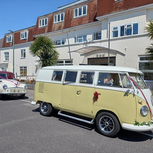 Sunny three‑star hotel entrance with palms and canopy, classic yellow VW camper and vintage wedding car parked outside.