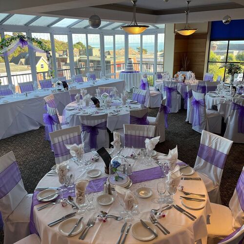 Light-filled banquet room set for a wedding with round tables, purple chair sashes, floral arch and distant sea view.