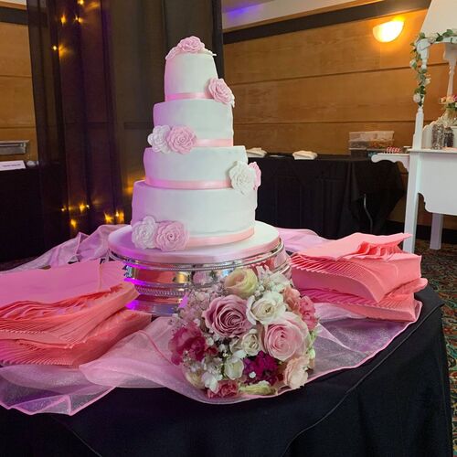 Five-tier white wedding cake with pink ribbons and sugar roses on dessert table with bouquet, napkins and candy cart