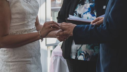 Bride and groom exchanging rings during intimate indoor wedding ceremony, bride in lace dress with beaded sash, officiant holding vows.