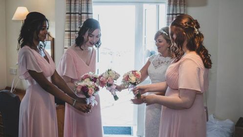 Bride and bridesmaids in pale pink dresses exchanging bouquets in a sunlit hotel suite getting-ready moment.