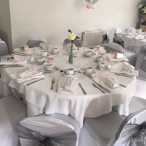 Round table set for a baby shower with white linens, teacups, favors, gray organza chair bows and single white rose.