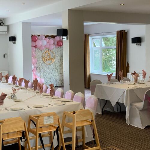 Bright dining room set for a baby shower with white tables, pink napkins, chair sashes and floral 'Oh Baby' balloon wall.