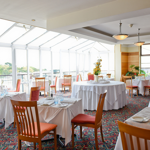 Bright conservatory hotel dining room with skylight windows, white-linen tables, floral centerpiece, wooden chairs and terrace views.