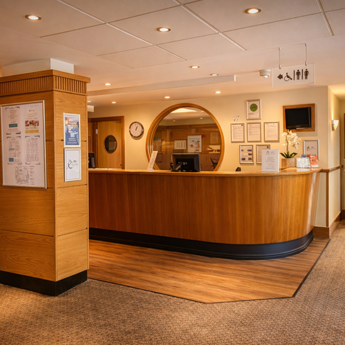 Warm wood reception desk with circular check-in window, orchid, accessible restroom signs; carpeted lobby leading to a set-up dining/meeting room.