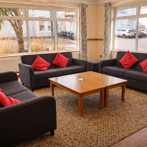 Cozy lounge with three dark sofas and red cushions around a wooden coffee table, large windows overlooking street parking.