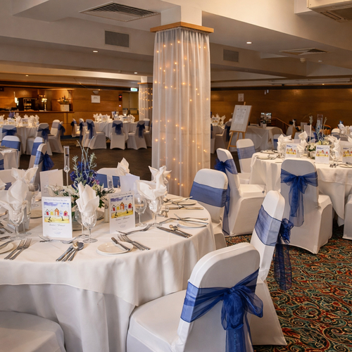 Elegant banquet hall set for wedding reception: round tables with white linens, navy-blue chair sashes, floral centerpieces and fairy-lit columns.