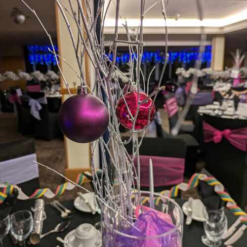 Silver-branch centerpiece with purple and magenta baubles over a dressed banquet table — black chairs with pink sashes, glassware and blue lights.