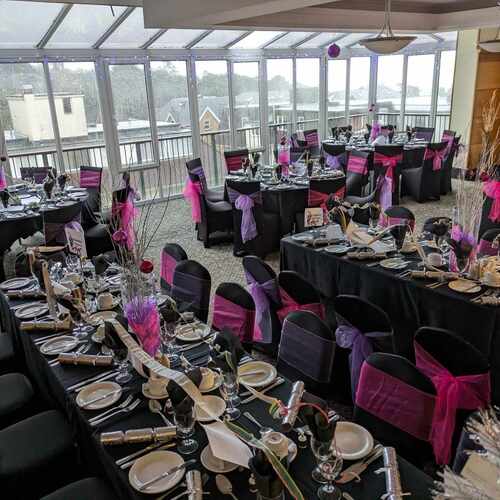 Glass-front banquet room with balcony, black-linen tables and pink and purple chair sashes, elegant place settings and festive centerpieces