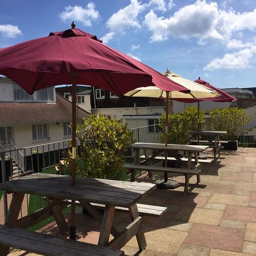 Rooftop terrace with wooden picnic tables, colorful parasols, shrubs and blue sky — al fresco seating for guests.