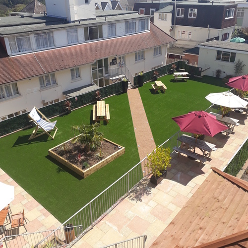 Sunny hotel courtyard with artificial lawn, picnic benches and umbrellas, deckchairs, raised planter and paved terrace with steps.