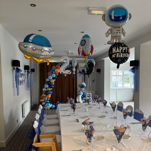 Private dining room set for a space-themed 2nd birthday: long white table with place settings, navy sashes, astronaut and rocket balloons.