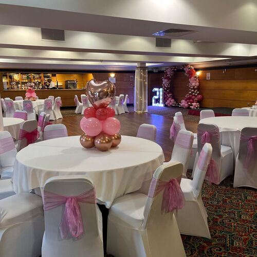 Banquet room set for a 21st: round tables with white linens, chairs tied with pink bows, heart balloon centerpieces, balloon arch, lit '21' and cake