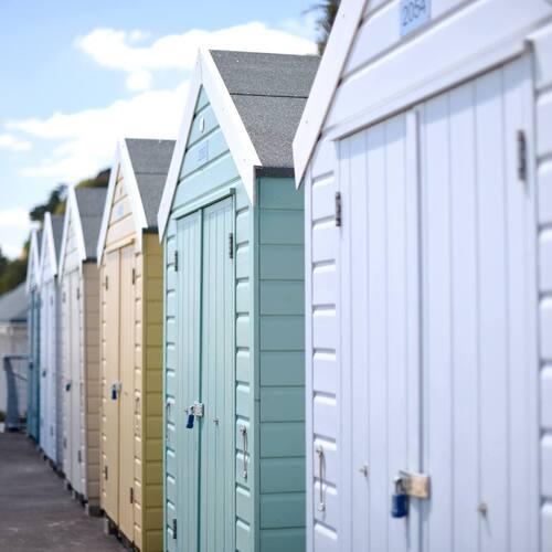Row of pastel beach huts with pitched roofs and padlocks lining a sunny seaside promenade, blurred beach and trees beyond.
