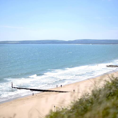 Sandy beachfront with gentle turquoise waters, wooden groynes and distant headland; sunlit shore with people walking.