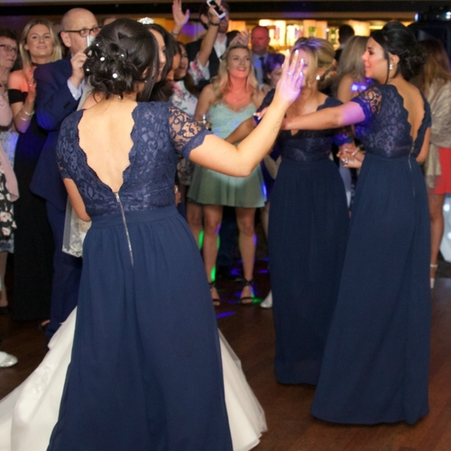 Bridesmaids in navy lace gowns dancing at lively hotel wedding reception, guests clapping around a wood dance floor.