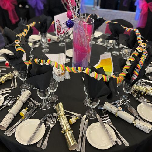 Banquet table set for a festive dinner with black linens, black napkins, place settings, party crackers, colorful streamers and pink centerpiece.