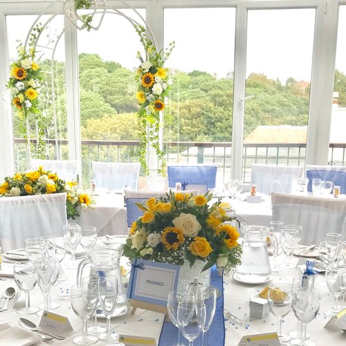 Hotel's sunlit glass-walled wedding reception with white linens, blue chair sashes, sunflower and yellow-rose bouquets, floral arch and treetop view