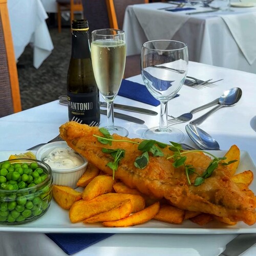 Crispy beer-battered fish with chunky chips, garden peas and tartare, paired with a chilled Prosecco flute on a white table.