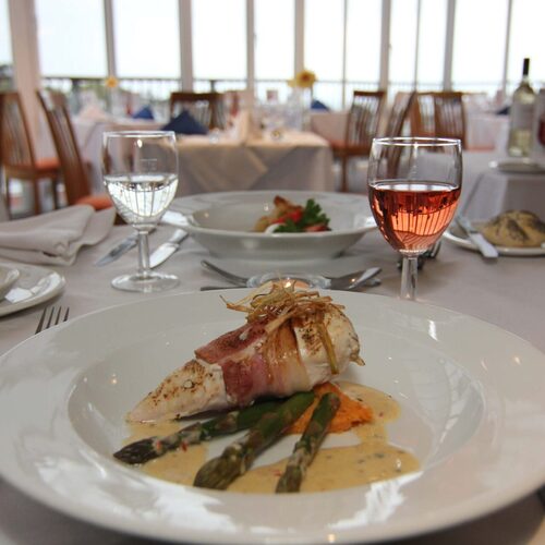 Fine-dining hotel restaurant with bacon-wrapped chicken, asparagus in cream sauce, rosé glass, white-linen table and bright terrace windows.