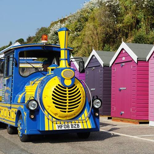 Bright blue-and-yellow tourist train beside pink and purple beach huts on a sunny seaside promenade for family rides