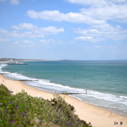 Sunny beachfront with sandy shore, turquoise sea and pier, clifftop promenade and hotels, families walking under blue sky.