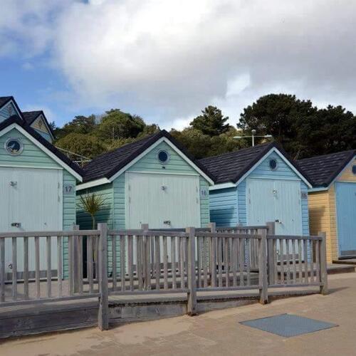 Row of pastel beach huts with private wooden decks on a sandy beachfront, coastal trees behind and a calm, overcast sky
