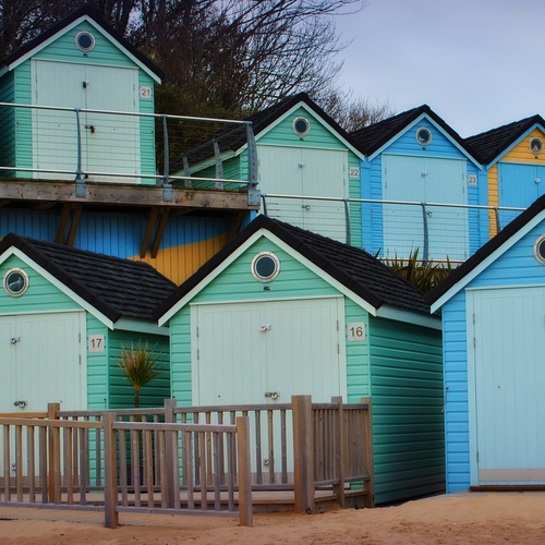 Rows of pastel beach huts with round porthole windows, numbered doors, a wooden boardwalk and sandy shoreline