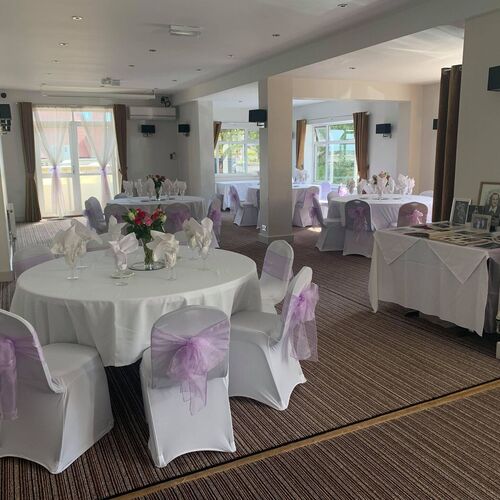 Bright hotel function room set for a wedding: round tables with white linens, lilac chair sashes, rose centerpieces and window light.