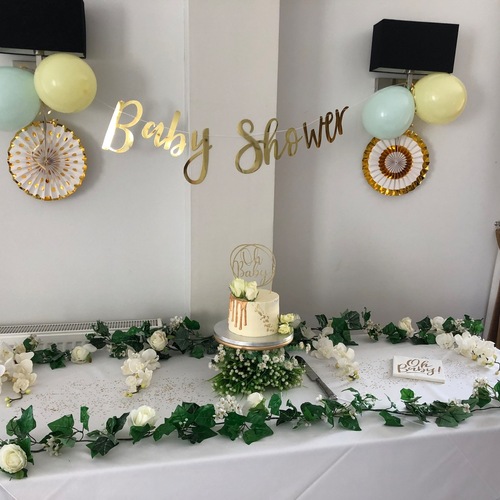 Elegant baby shower table with white cake and gold drip topper, pastel balloons, 'Baby Shower' gold banner and garlands.