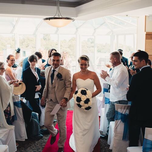 Bride in strapless gown and groom in tan suit walk down red aisle in sunlit room as guests applaud, chairs with blue sashes.