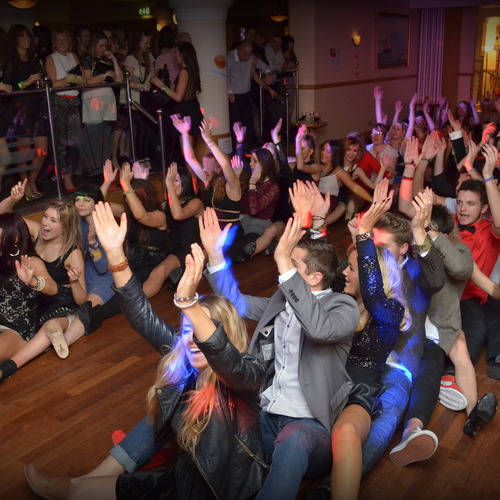 Lively hotel ballroom party on wooden dance floor, guests in evening wear in a conga line with hands raised.