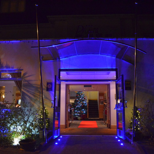 Hotel entrance with glass canopy and blue uplighting, open doors to a red carpet and decorated Christmas tree in the lobby.