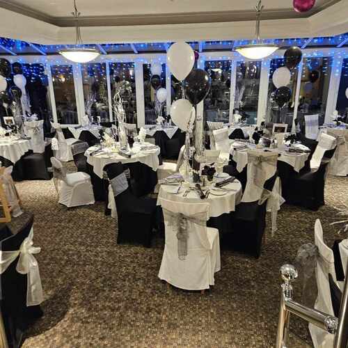 Banquet room set for celebration with black & white chair covers, round tables, white and black balloons and blue fairy lights.