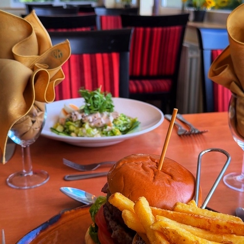 Gourmet burger with tomato and lettuce, basket of crispy fries and coleslaw on a wooden table in bright, cozy dining room.