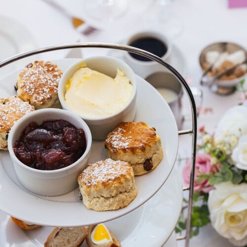 Three-tier tea stand with freshly baked scones, clotted cream and fruit jam, set on a white linen table next to floral centerpiece.