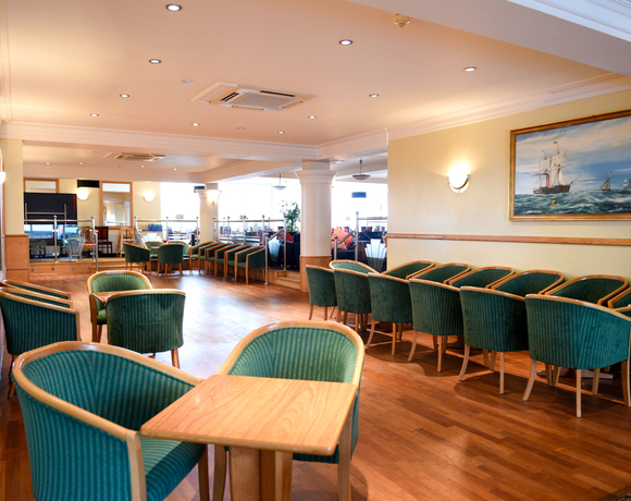 Spacious hotel lounge with rows of green upholstered chairs, wooden tables, warm lighting and a nautical painting.